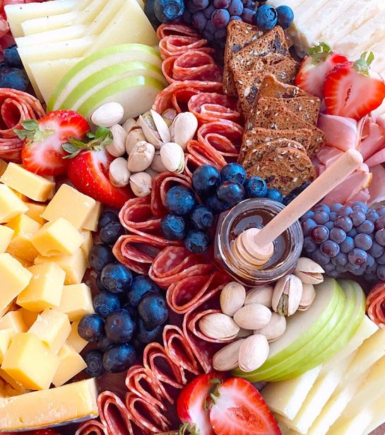 close up of charcuterie board with blueberries, strawberries, apples, honey, cheese, meats, and crackers